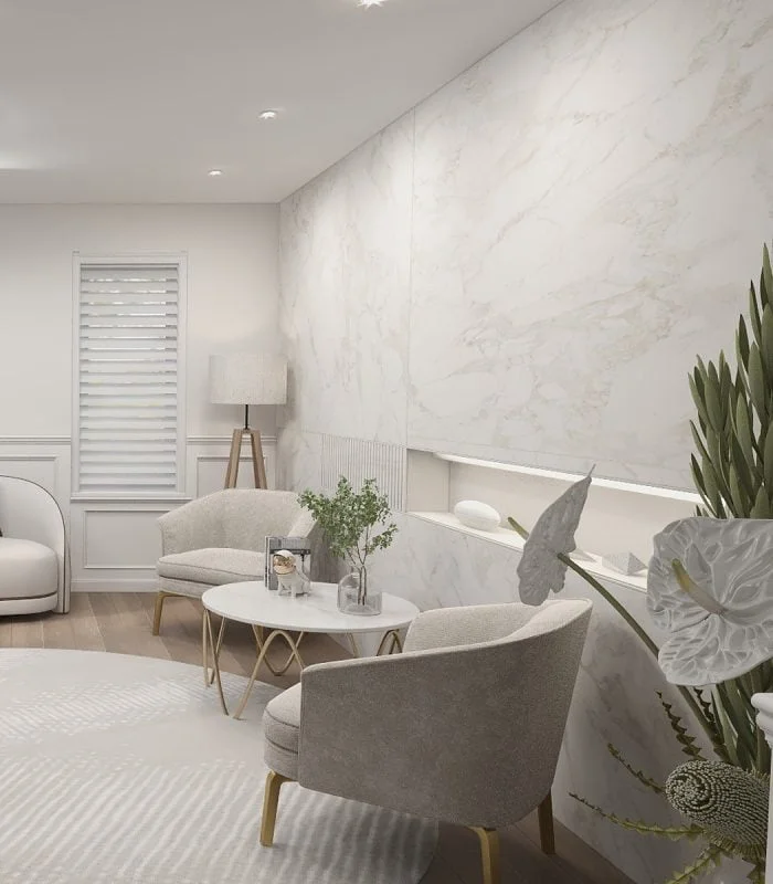İç mimarın tarzıyla tasarlanmış, Premium açık gri sandalyeler, yuvarlak masa, ayaklı lamba ve dekoratif bitkilerle donatılmış modern bir oturma odası. Mermer desenli duvarlar, panjurlu küçük pencereyi tamamlayarak, özel bir villanın zarafetini yansıtıyor.